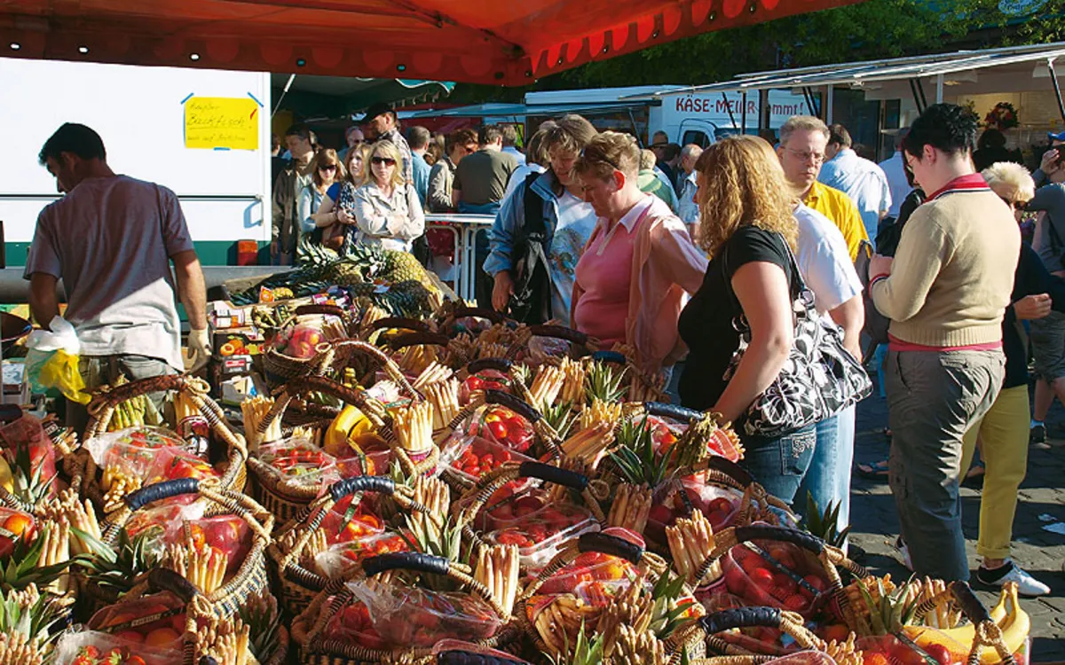 Menschen auf einem belebten Markt mit Körben voller Obst und Gemüsesorten unter einem roten Zelt.