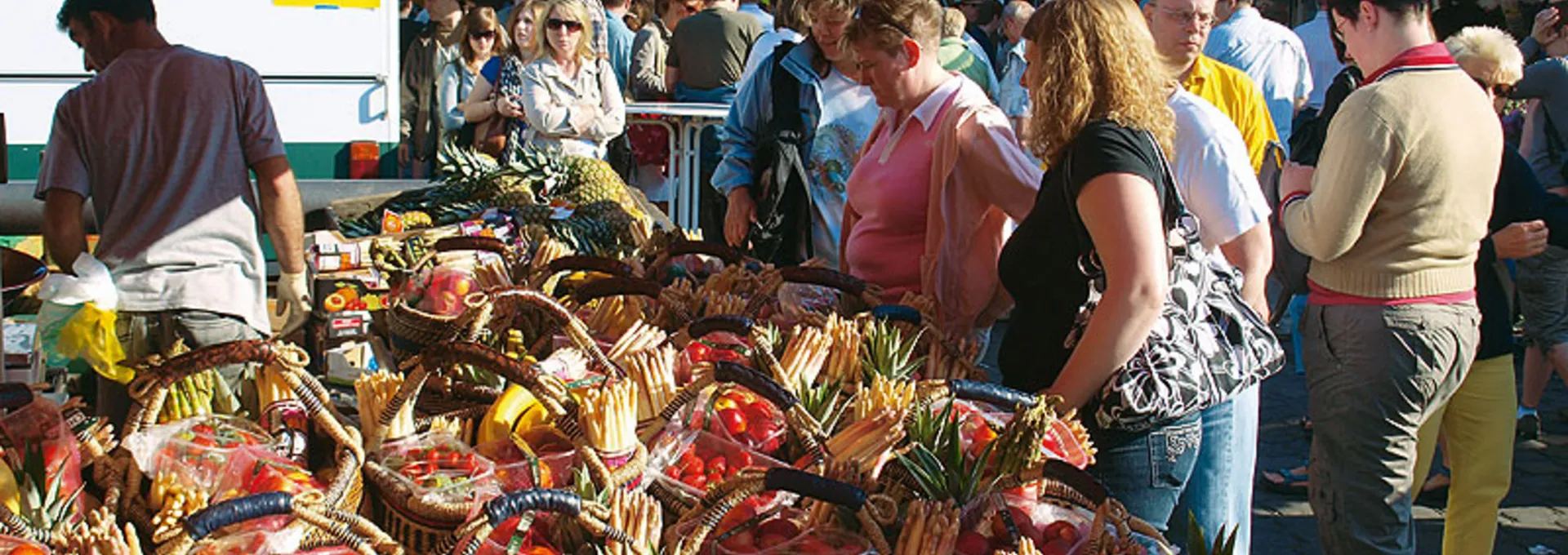 Menschen auf einem belebten Markt mit Körben voller Obst und Gemüsesorten unter einem roten Zelt.