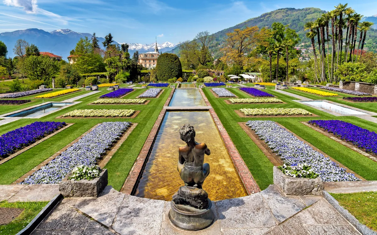 Garten mit Blumenbeeten, Brunnen und Bergen im Hintergrund.
