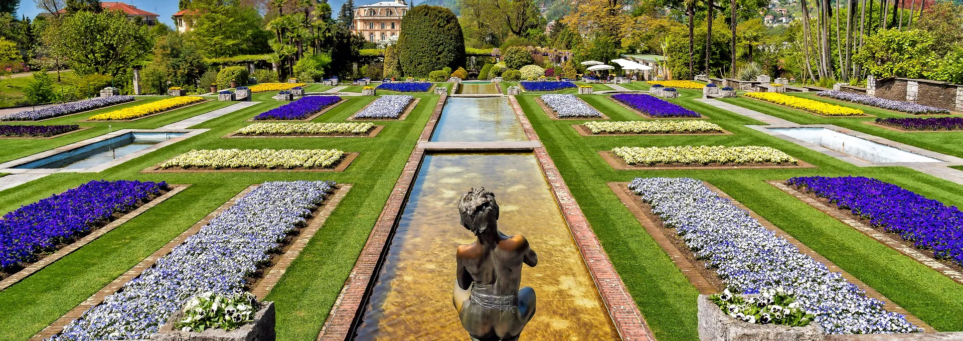 © EleSi - stock.adobe.com Garten mit Blumenbeeten, Brunnen und Bergen im Hintergrund.