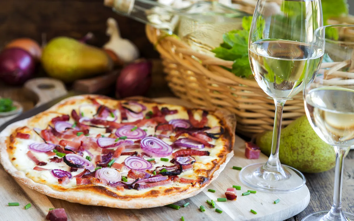 Pizza mit Zwiebeln und Speck neben Weißwein und einem Korb mit Birnen.