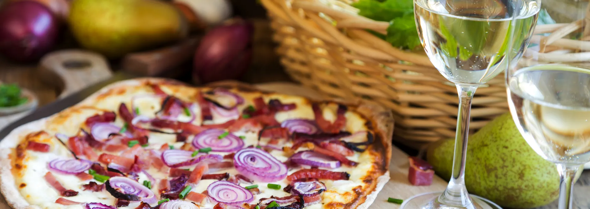 Pizza mit Zwiebeln und Speck neben Weißwein und einem Korb mit Birnen.