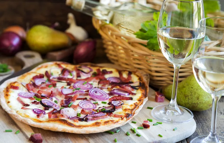 Pizza mit Zwiebeln und Speck neben Weißwein und einem Korb mit Birnen.