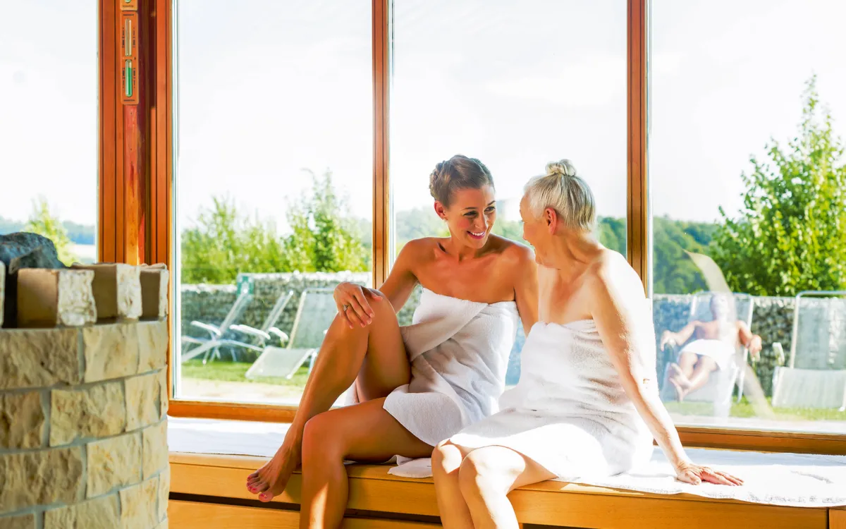 Zwei Frauen in Handtüchern sitzen lächelnd in einer Sauna mit Blick ins Freie.