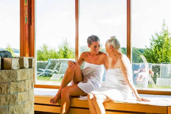 Zwei Frauen in Handtüchern sitzen lächelnd in einer Sauna mit Blick ins Freie.