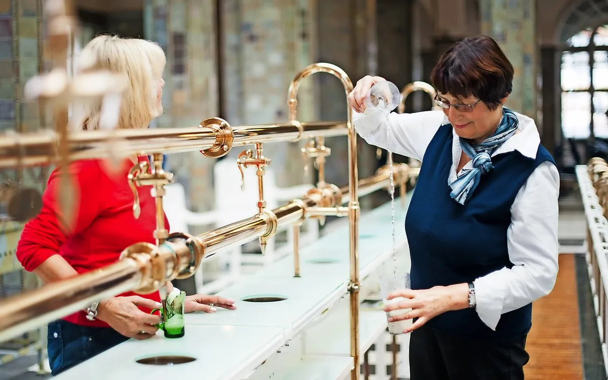 Zwei Frauen beim Ausschank von Heilwasser an einer metallenen Wasserleitung.