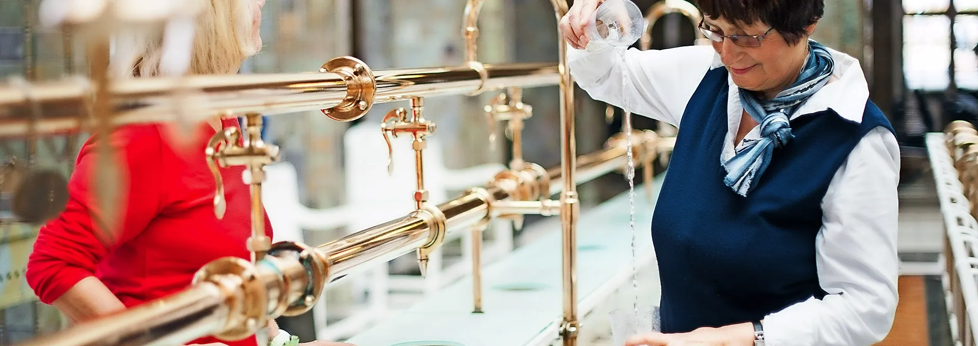 Foto: Heji Shin Zwei Frauen beim Ausschank von Heilwasser an einer metallenen Wasserleitung.