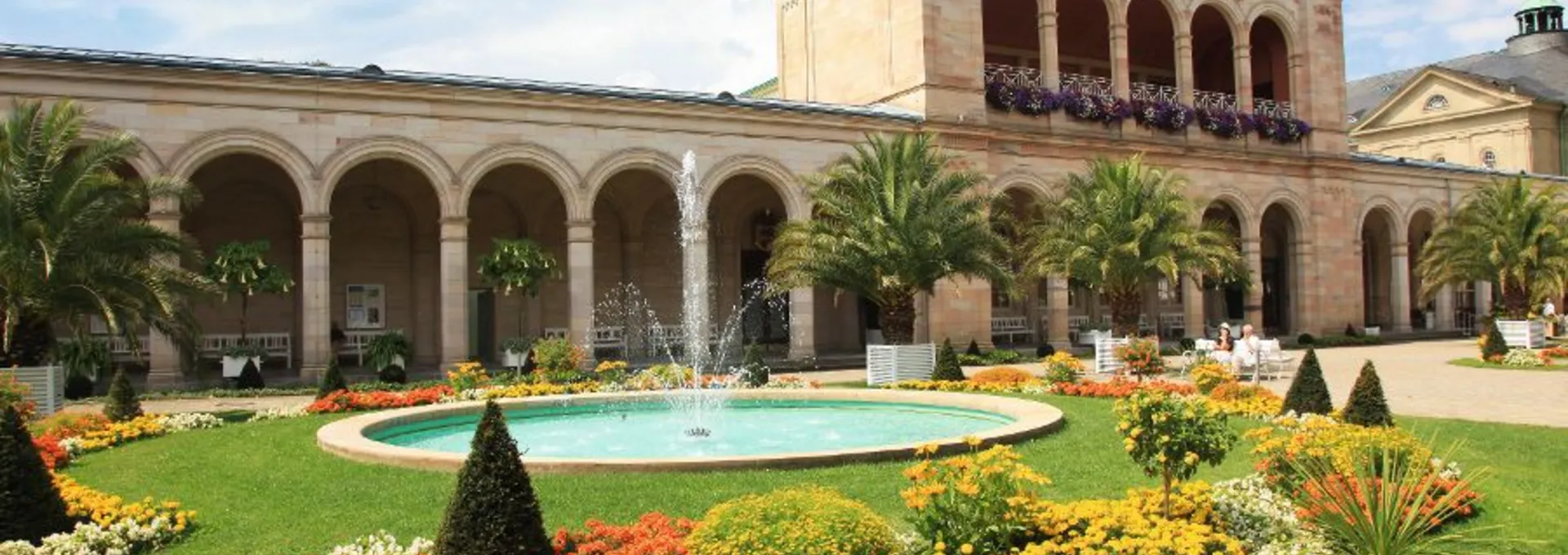 © Bayer. Staatsbad Bad Kissingen GmbH Arkaden mit Springbrunnen und blühenden Gärten unter blauem Himmel in Bad Kissingen.